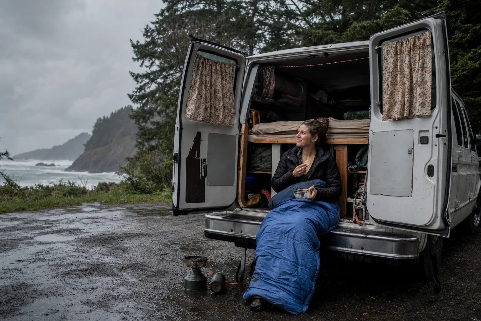 Elena eats crackers from her sleeping bag at the open back of her van on the rainy Pacific coast
