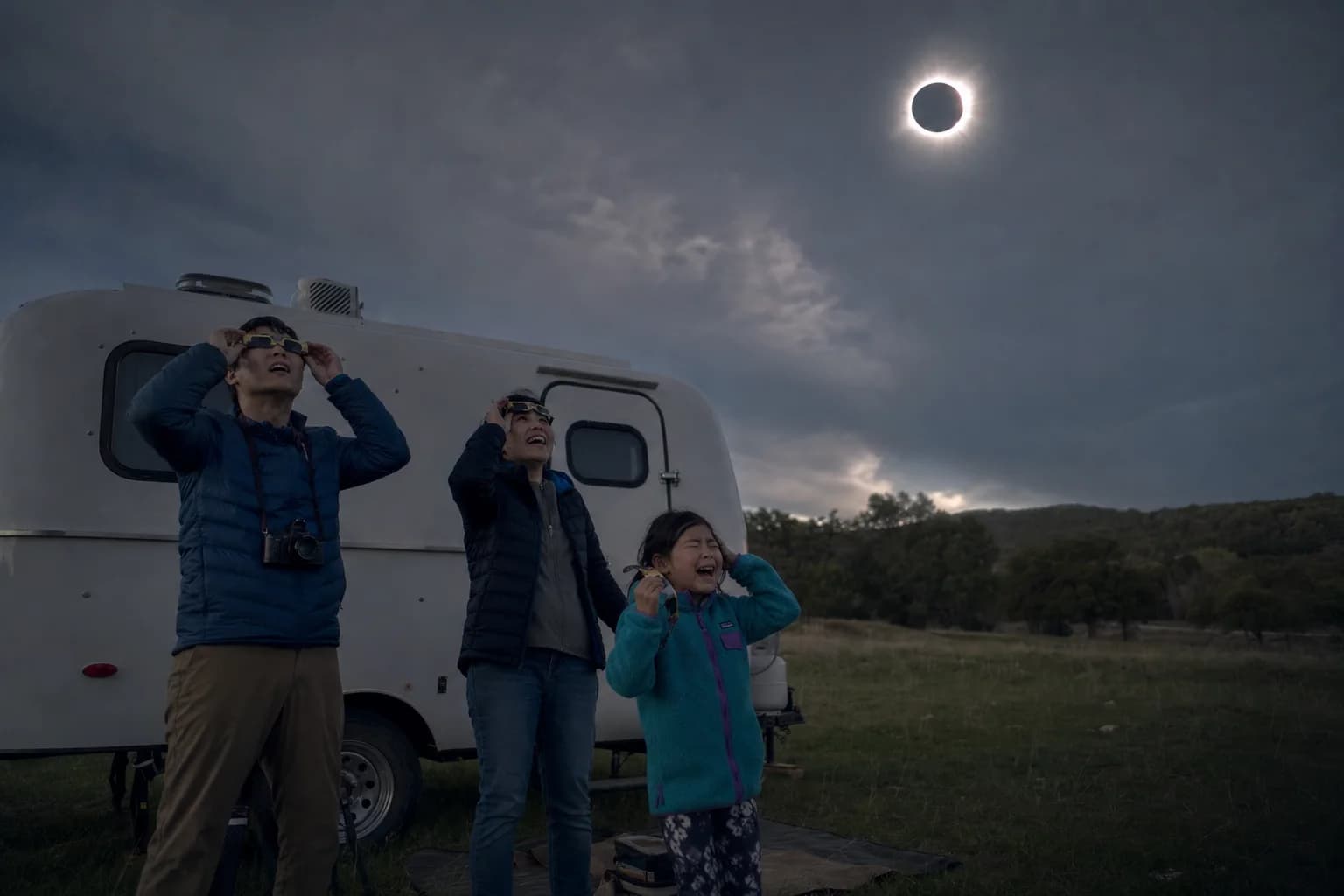Naomi, Kenji, and Emi watch the total solar eclipse beside their tiny fiberglass trailer in Texas
